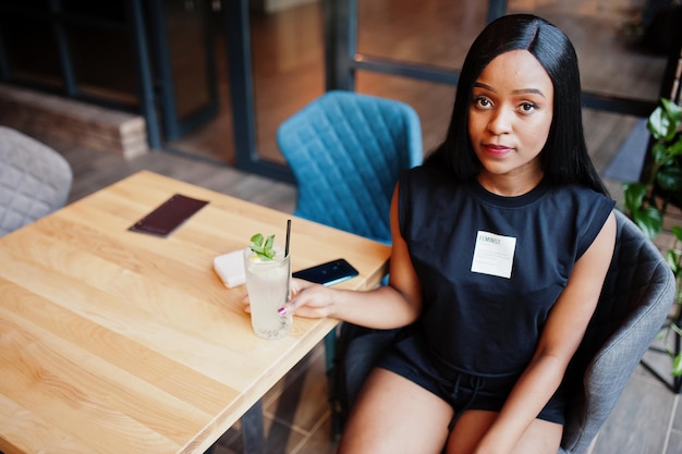 Modische feministische Afroamerikanerin in schwarzem T-Shirt und Shorts, posiert im Restaurant mit Limonadenglas.