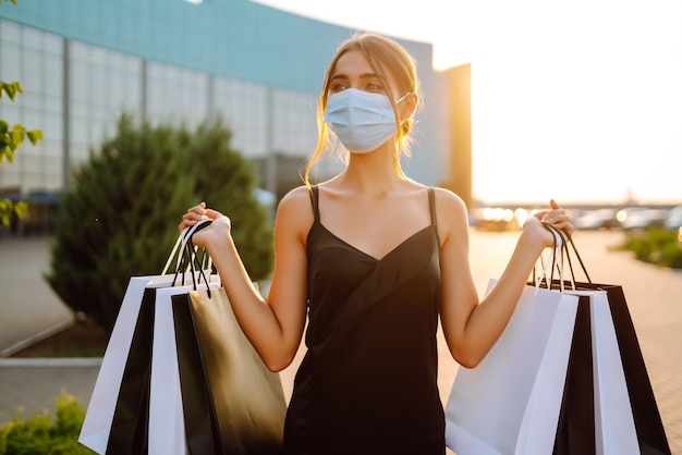 Modische Frau, die medizinische Schutzmaske mit Einkaufstaschen bei Sonnenuntergang trägt.