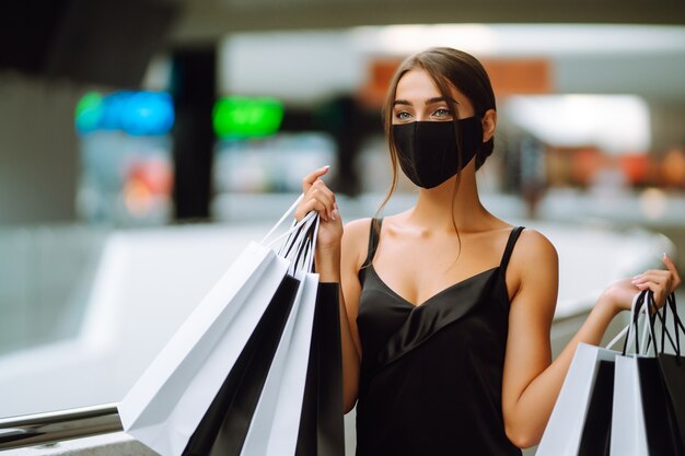 Modische Frau, die medizinische Schutzmaske mit Einkaufstaschen im Einkaufszentrum trägt.