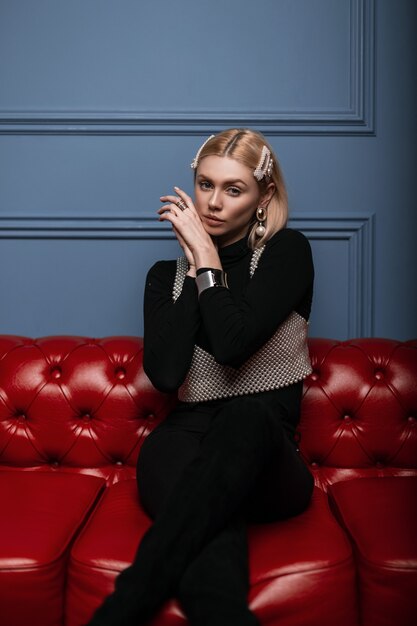 Foto modische glamouröse junge blonde frau in schwarzer, stilvoller kleidung in einem eleganten perlenoberteil mit einem vintage-haar mit eleganten perlenclips, das auf einem roten ledersofa posiert