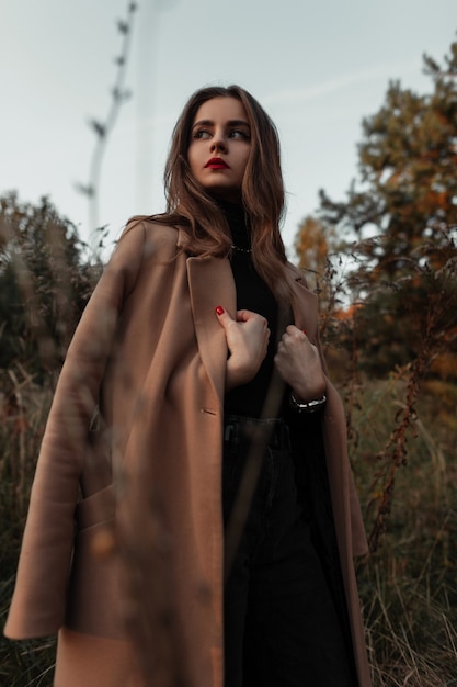 Modische Herbstmodellfrau mit roten Lippen in einem beigefarbenen Mantel des Modeherbstes im Wald in der Natur. Outfit-Look im Vogue-Stil