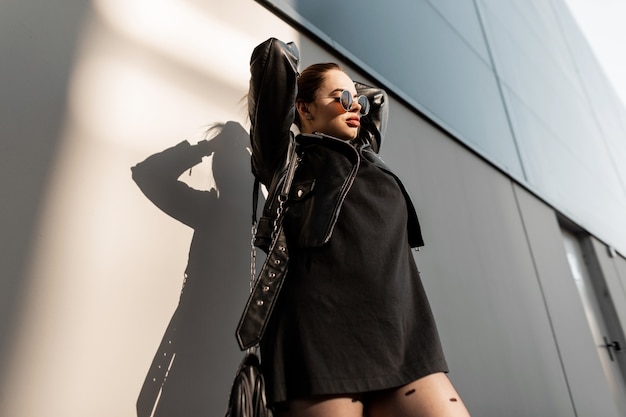 Modische Hipster-Frau mit runder Vintage-Sonnenbrille in stilvoller schwarzer Kleidung mit Lederjacke und Kleid mit Handtasche steht in der Nähe der Wand in der Stadt im Sonnenlicht