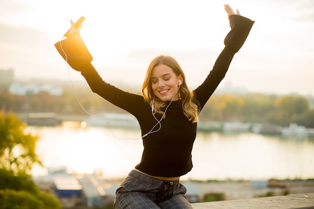 Modische hörende Musik der jungen Frau vom Smartphone im Freien bei Sonnenuntergang