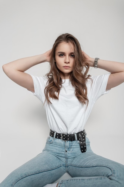 Modische hübsche junge Frau mit lockiger Frisur im trendigen weißen T-Shirt mit Vintage-Blue-Jeans sitzt im Studio