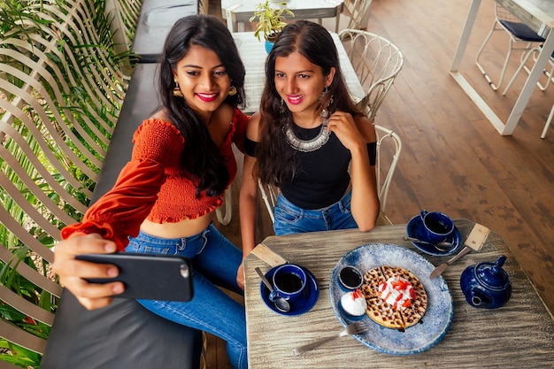 Modische indische Freunde Paar von zwei Frauen, die Selfie-Handy machenFreunde lang erwartetes Treffen mit Klatschstudenten, die Pfannkuchen essen und Masala-Tee nach dem Einkaufen im Sommer instahram-Lebensstil essen