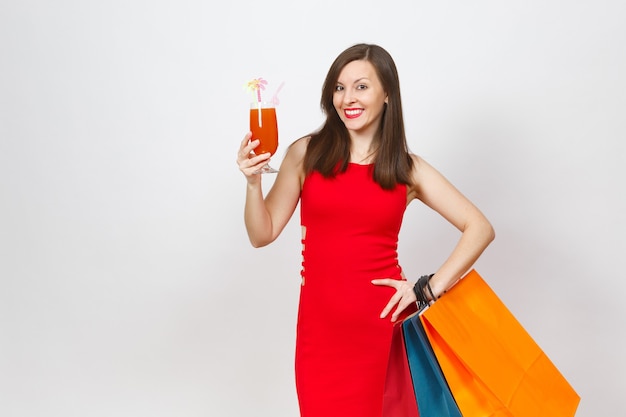 Modische junge Frau des attraktiven Glamours im roten Kleid, das ein Glas Getränkcocktail hält, mehrfarbige Pakete mit Einkäufen nach dem Einkaufen lokalisiert auf weißem Hintergrund. Kopieren Sie Platz für Werbung.