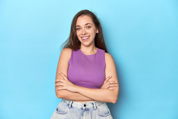 Modische junge Frau in einem lila Top auf blauem Hintergrund, die sich zuversichtlich fühlt und die Arme kreuzt