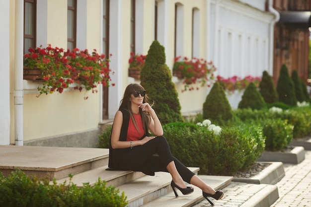 Modische junge Frau in trendiger Sonnenbrille und roter Bluse, die am sonnigen Sommertag auf der Treppe auf der Stadtstraße sitzt.