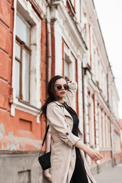 Modische junge Frau in Vintage-Sonnenbrille in stilvollem Trenchcoat in schwarzer Kleidung mit trendiger Ledertasche in der Nähe des alten Gebäudes in der Stadt. Wunderschönes Mädchenmodell geht im Freien. Jugend weiblicher urbaner Stil.