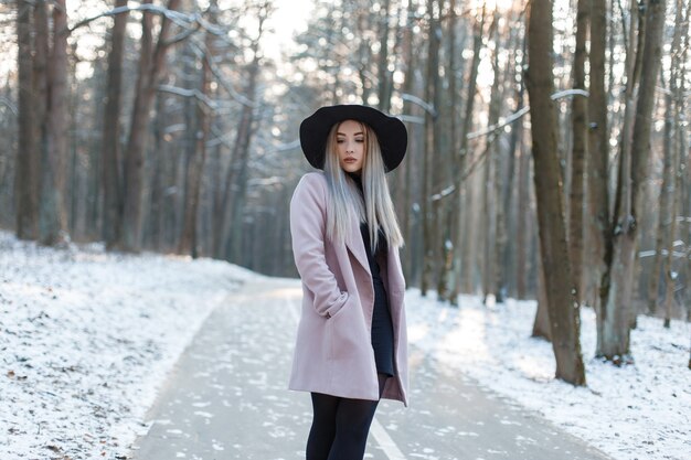 Modische junge Fraublondine in einem stilvollen glamourösen Mantel in einem schwarzen Kleid in einem stilvollen schwarzen Hut steht in einem verschneiten Winterwald an einem sonnigen Tag. Attraktives Mädchen auf einem Spaziergang. Moderne Damenmode