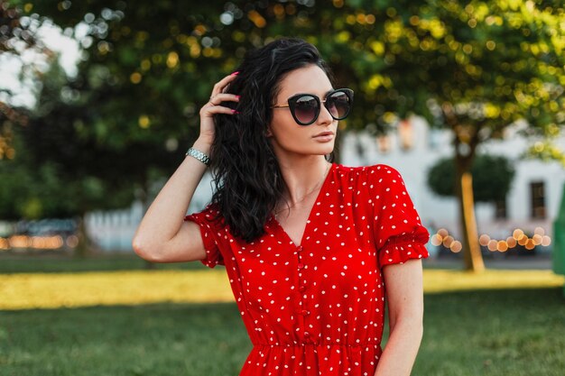 Modische lockige Frau mit Sonnenbrille und modischem rotem Kleid, die im Park spazieren geht