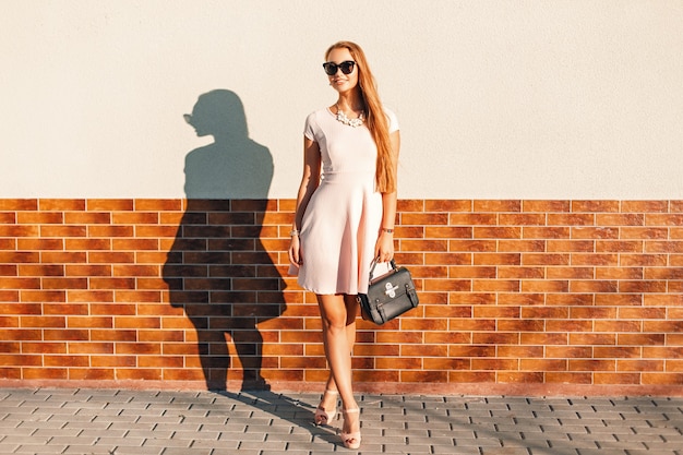 Modische schöne Frau in der Sonnenbrille, rosa Kleid, das Tasche nahe Ziegelmauer hält