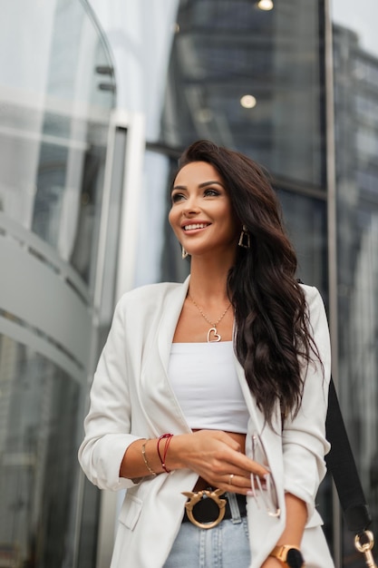 Modische, schöne Geschäftsfrau mit Lächeln in stylischem weißem Blazer-Oberteil und Jeans mit Tasche geht spazieren und genießt in der modernen Stadt Schönheitsmädchen im stilvollen Freizeitoutfit-Look