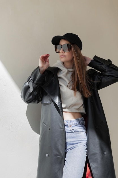 Modische, schöne Hipster-Straßenfrau mit cooler Sonnenbrille und schwarzer Mütze, die modische Freizeitkleidung mit Ledermantel und Hoodie trägt, steht in der Nähe einer Mauer in der Stadt