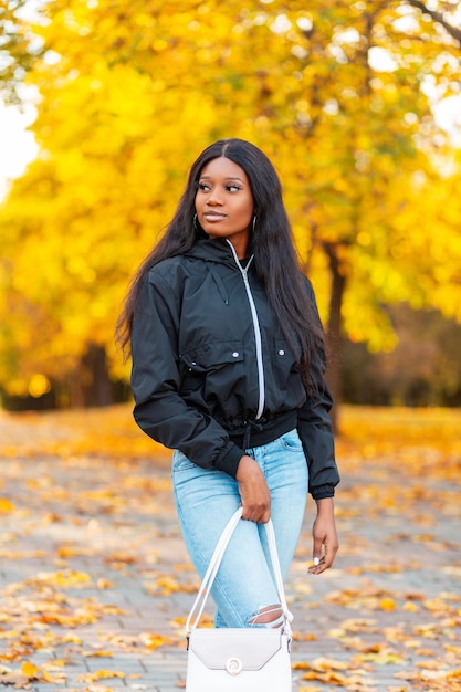 Modische schöne junge afroamerikanische Frau in stylischer, lässiger schwarzer Jacke mit Blue Jeans und Blue Jeans mit Handtasche geht in einem Herbstpark mit buntem goldenem Laub