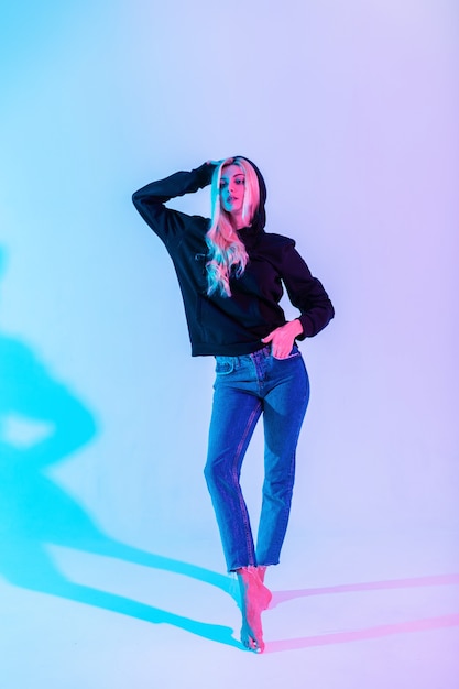 Modische schöne junge Frau mit schlankem Körper mit stylischem schwarzen Hoodie und blauen Jeans auf mehrfarbigem rosa Neonhintergrund im Studio