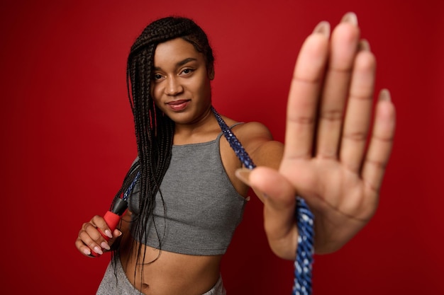 Modische, sportliche, fröhliche Afrikanerin mit stylischen Dreadlocks, die vor einem roten Hintergrund mit einem Springseil um den Hals posiert, gestikuliert, ein STOP-Schild zeigt und in die Kamera schaut. Platz kopieren