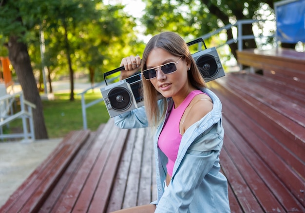 Modische, stilvolle blonde Kaukasierin mit Sonnenbrille in Jugendsommerkleidung, die ein Retro-Tonbandgerät hält