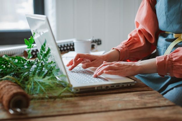 Modische, stilvolle Hipster-Floristin in einer Arbeitsschürze, die auf einem Laptop arbeitet und auf ein Smartphone blickt