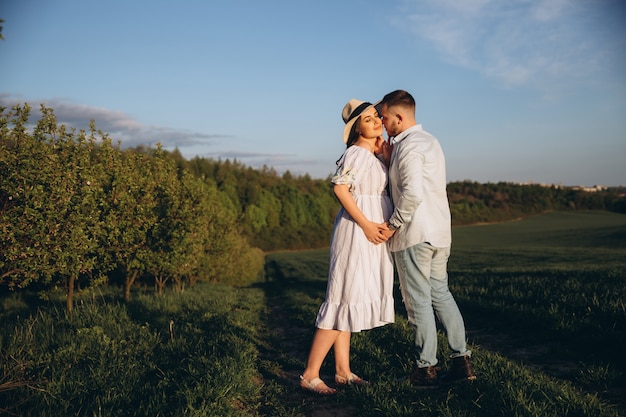 Modische und stilvolle glückliche schwangere Frau und ihr Ehemann kleideten einen pastellweißen und blauen Ton auf dem Feld auf dem Sonnenuntergang