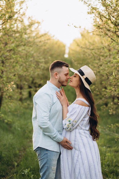 Modische und stilvolle glückliche schwangere Frau und ihr Ehemann kleideten einen pastellweißen und blauen Ton im Garten auf dem Sonnenuntergang an