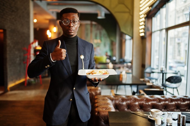 Modischer afrikanisch-amerikanischer Mann in Anzug und Brille steht im Café mit Teller Salat in der Hand und zeigt Daumen nach oben