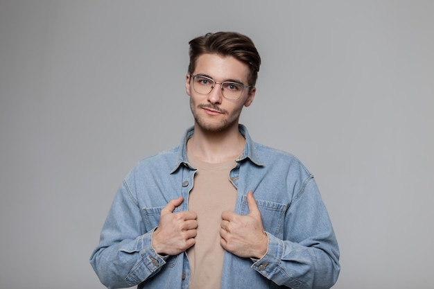 Modischer amerikanischer Hipster-Mann mit Vintage-Sonnenbrille, Jeanshemd und beigem T-Shirt auf grauem Hintergrund im Studio Männliches Porträt