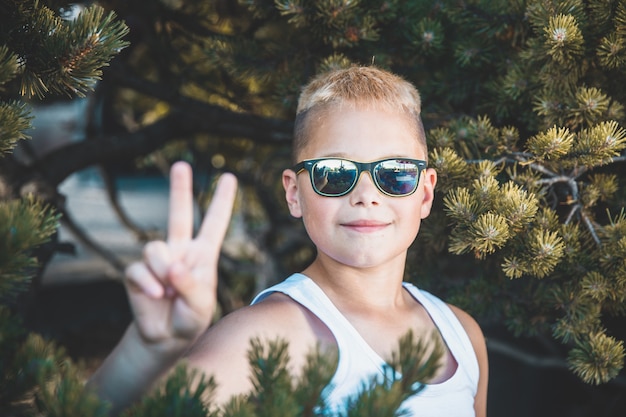 Modischer blonder Junge mit Sonnenbrille zeigt zwei Finger.