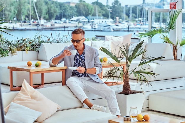 Modischer erfolgreicher Mann mit stylischem Haar in moderner, eleganter Kleidung hält eine Tasse Kaffee, während er auf einem Sofa im Café im Freien vor dem Hintergrund des Stadtkais sitzt.