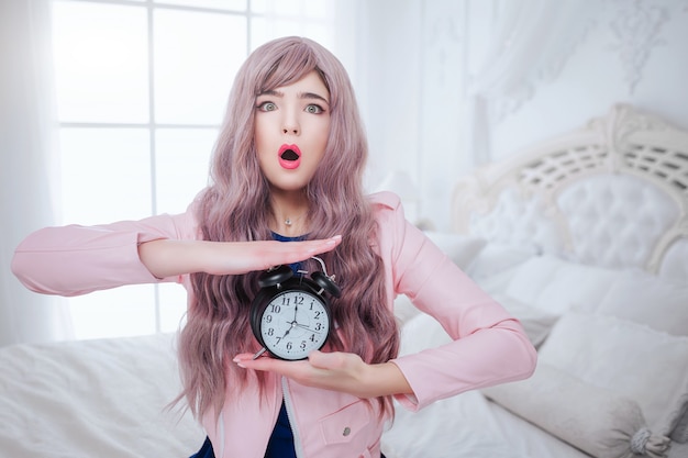 Modischer Freak. Glamour emotionale schöne Frau mit langen lila Haaren hält schwarze Uhr, während im weißen Schlafzimmer sitzen. Schönheits- und Modekonzept.