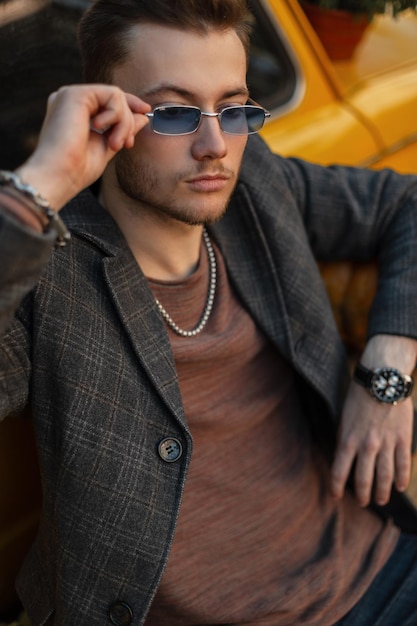 Modischer Geschäftsmann-Hipster-Typ in Freizeitkleidung mit Modemantel und Luxusuhr trägt eine stylische Sonnenbrille und sitzt in der Nähe eines gelben Retro-Autos