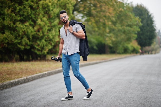 Modischer großer arabischer Bartmann trägt Hemdjeans und Sonnenbrille, die mit Regenschirm und Mantel zur Hand im Park spazieren gehen