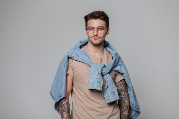 Modischer, gutaussehender Hipster-Typ mit Haaren und Tattoos auf den Armen, der stylische Freizeitkleidung mit einem Jeanshemd auf den Schultern, einem beigen T-Shirt und einer Vintage-Brille im Studio trägt