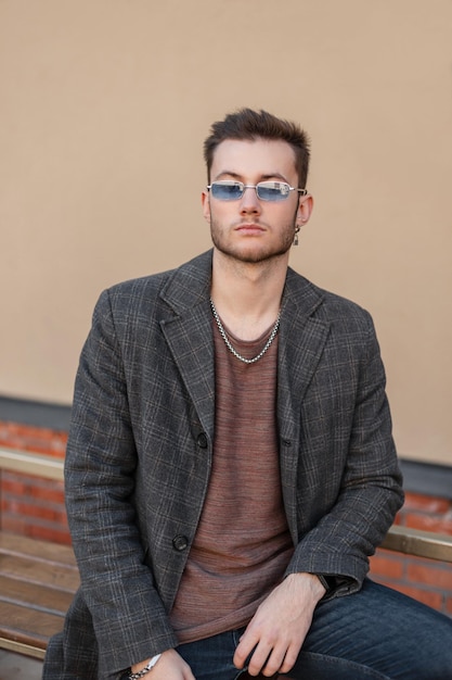 Modischer Hipster mit stylischer blauer Sonnenbrille in modischem Mantel und T-Shirt sitzt auf der Straße in der Nähe einer Mauer