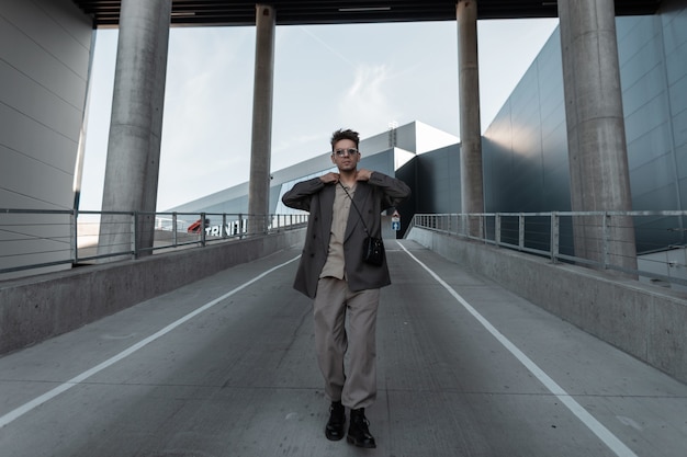 Modischer junger Geschäftsmann mit stylischer Sonnenbrille in einem trendigen grauen Anzug mit Handtasche geht in der Stadt auf die Straße