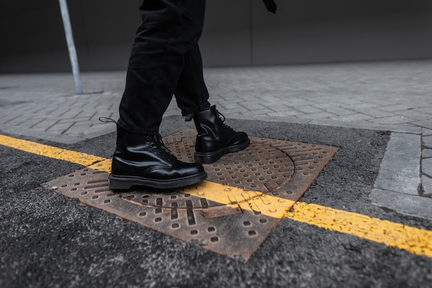 Modischer junger Mann in schwarzen Lederstiefeln in trendigen Jeans steht auf einer eisernen Luke in der Stadt. Nahaufnahme von männlichen Beinen in stilvollen saisonalen Schuhen. Streetstyle mit gelber Linie. Jugendmode.