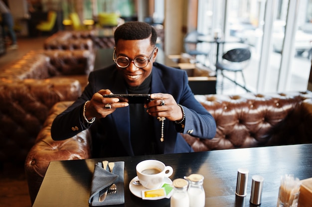 Modischer Mann im Anzug und in den Gläsern, die am Café sitzen und Kaffee am Telefon schnappen