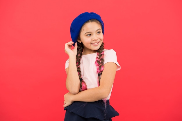 Modisches Baskenmütze-Accessoire Teenagermode Französisches Modeattribut Kind kleines Mädchen glücklich lächelndes Baby Kind kleines süßes Modemädchen, das mit langen Zöpfen und Hut auf rotem Hintergrund posiert Modemädchen