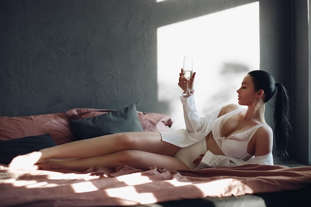Modisches brünettes Mädchen in Dessous, das auf dem Bett liegt und mit Champagner einen luxuriösen Lebensstil genießt