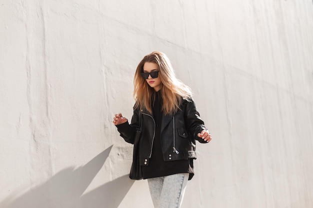 Modisches Hipster-Frauenmodell mit Sonnenbrille in trendigem Lederjacken-Hoodie und Tasche geht auf der Straße in der Nähe einer grauen Wand spazieren