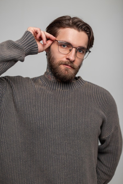 Modisches, hübsches, männliches Porträt eines hübschen, stilvollen, hipster, erfolgreichen Mannes mit Bart und Frisur in einem modischen, vintage gestrickten Pullover passt seine Brille im Studio an