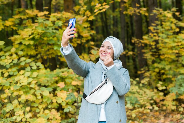 Modisches junges muslimisches asiatisches Mädchen im Hijab, das draußen im Herbstpark ein Selfie auf dem Smartphone macht