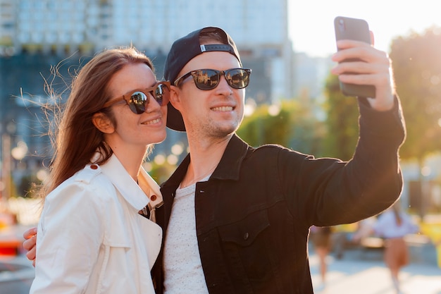 Modisches Paar, das Selfie mit dem Handy macht. Mann und Frau mit Sonnenbrille treffen Freunde in der Stadt Datumsfoto auf einem Smartphone gegen den Sonnenuntergang.