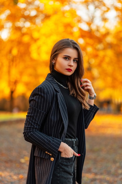 Modisches Porträt einer schönen eleganten Frau mit roten Lippen in einem stylischen Blazer mit Pullover auf dem Hintergrund eines erstaunlichen Herbstparks mit gelbem Herbstlaub