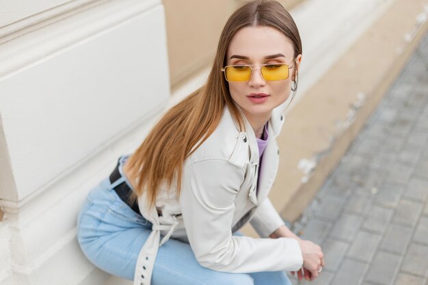 Modisches, schönes Frauen-Hipster-Modell mit gelber Sonnenbrille in einer trendigen weißen Lederjacke und Blue Jeans sitzt in der Nähe eines alten Gebäudes an der Wand