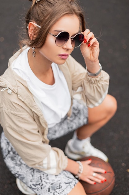 Modisches, schönes Hipster-Mädchen in Retro-Sportbekleidung mit Windjacke, T-Shirt-Leggings und runder Vintage-Sonnenbrille sitzt mit dem Ball auf dem Basketballplatz