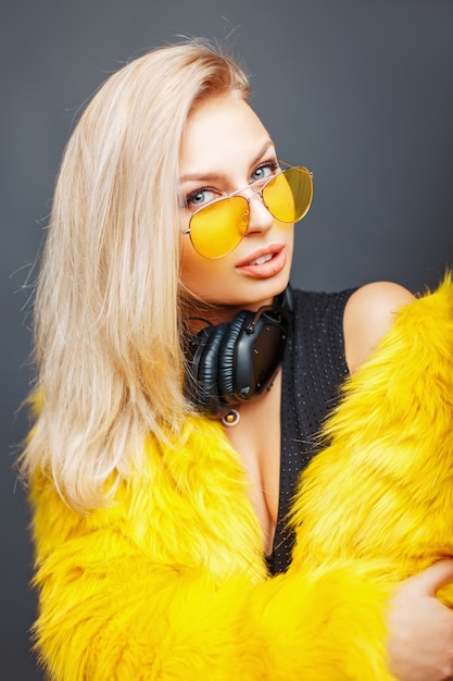 Modisches schönes junges DJ-Mädchen mit Kopfhörern in der stilvollen Brille und einem modischen gelben Pelzmantel auf Grau