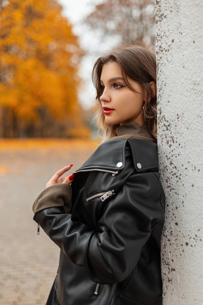 Modisches schönes Mädchen mit roten Lippen in einer modischen schwarzen Lederjacke steht in der Nähe einer weißen Wand in einem Herbstpark