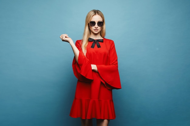 Modisches und sexy blondes Modellmädchen in der stilvollen Sonnenbrille und im glamourösen kurzen roten Kleid mit schwarzer Schleife, isoliert