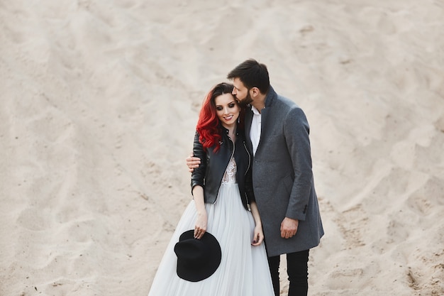Modisches und stilvolles Paar, sexy Model Girl mit roten Haaren und hellem Make-up, im Spitzenkleid und in stilvoller Lederjacke und hübschen bärtigen Männern im trendigen grauen Mantel, die in der Wüste posieren